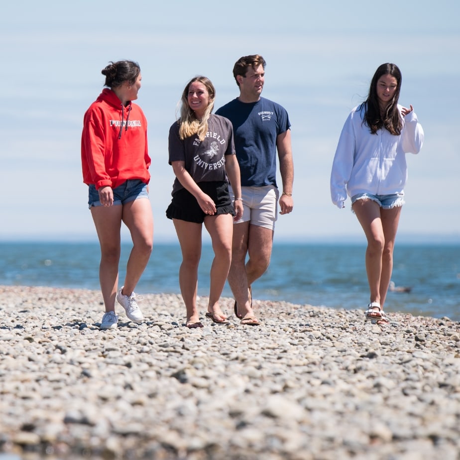 Four students walk side by side on the beach, surrounded by sand and waves, enjoying a sunny day.